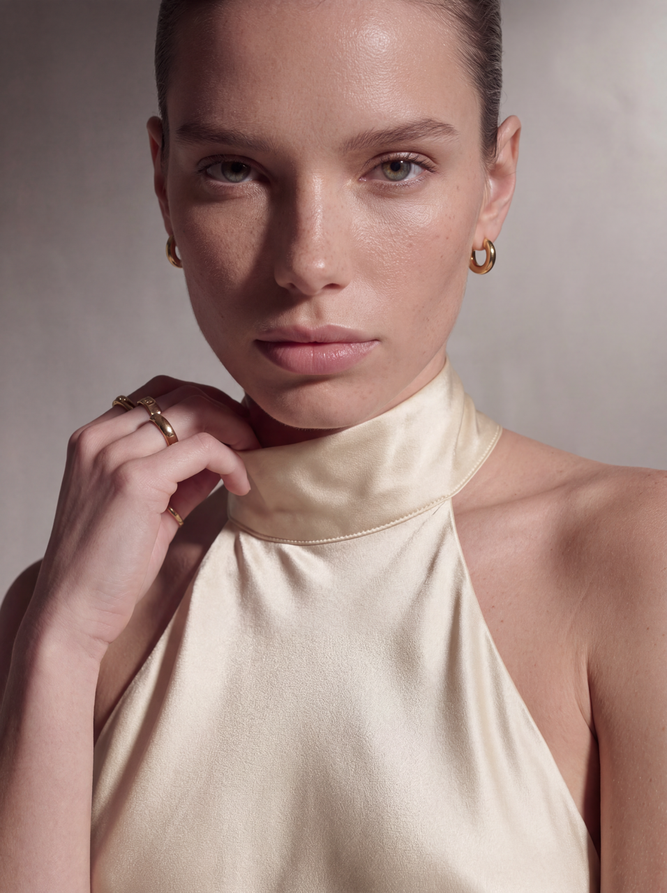 A close-up portrait of a model wearing a cream satin halter top and gold hoop earrings against a neutral beige background.