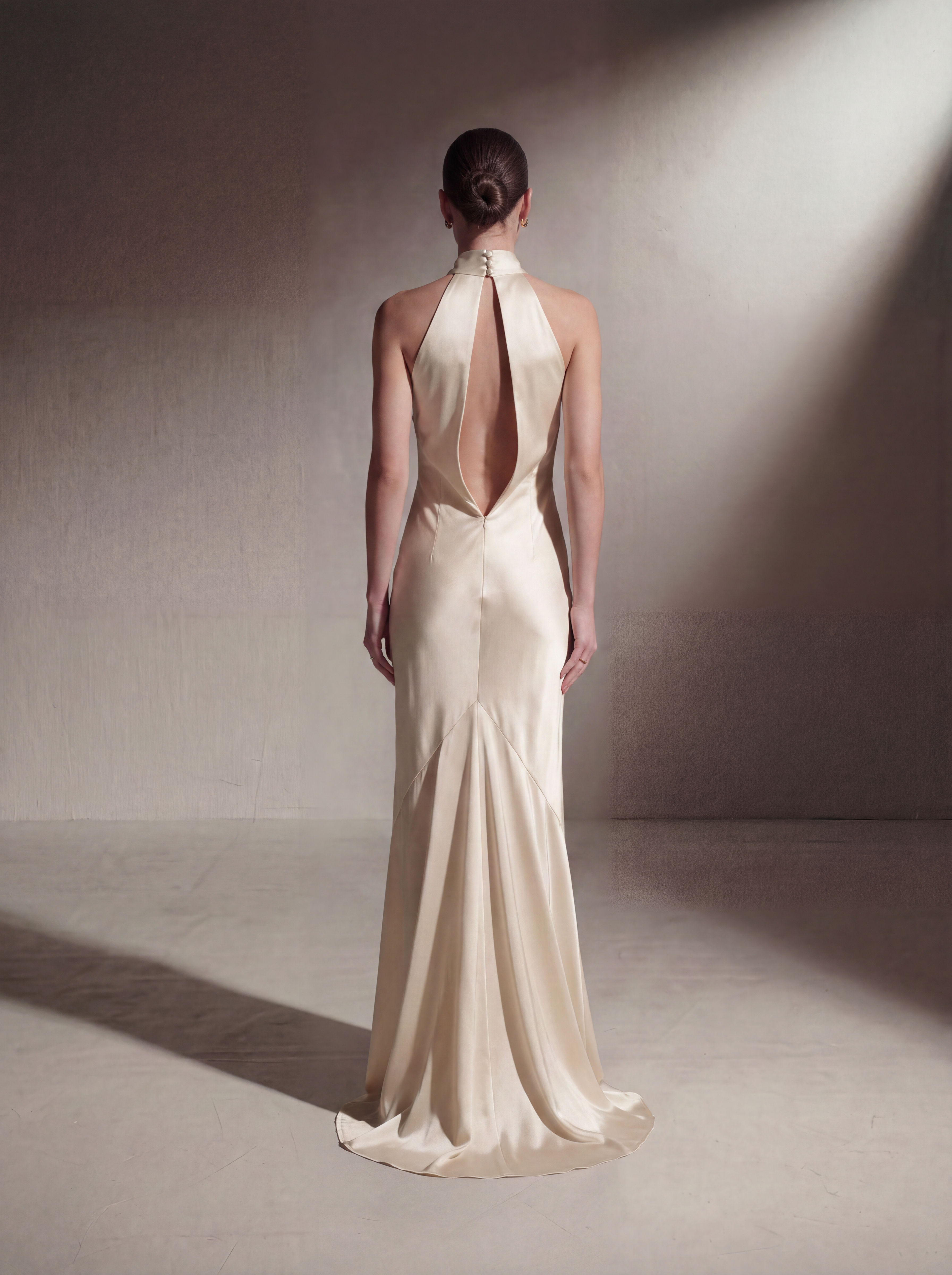 A model viewed from behind wearing a floor-length cream satin gown with a halter-style open back, standing in a studio with dramatic side lighting.