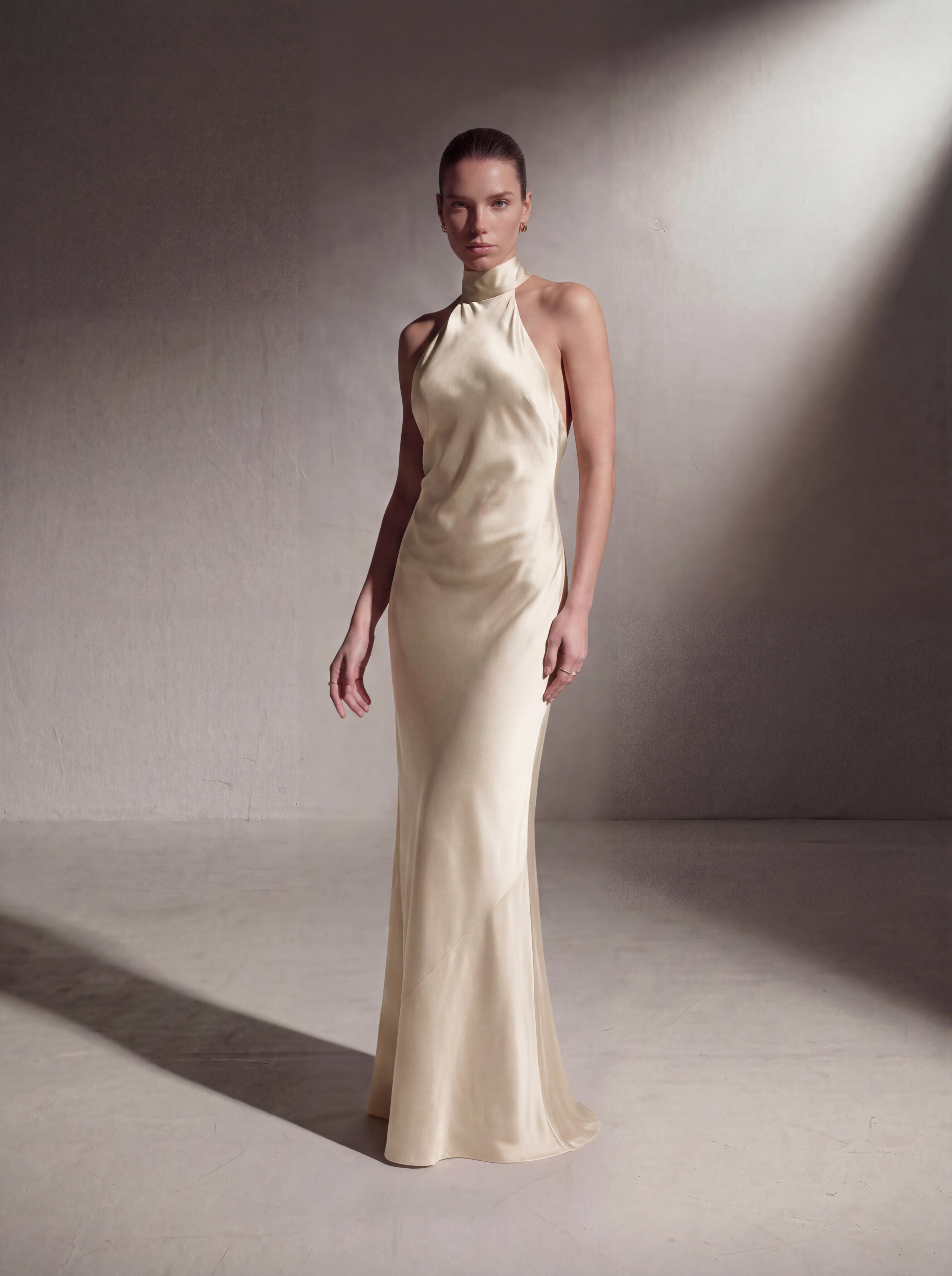 A model stands in a studio wearing a floor-length cream satin gown with a high halter-style neckline and a fitted silhouette. The background is a textured neutral wall and floor with dramatic directional lighting casting shadows.